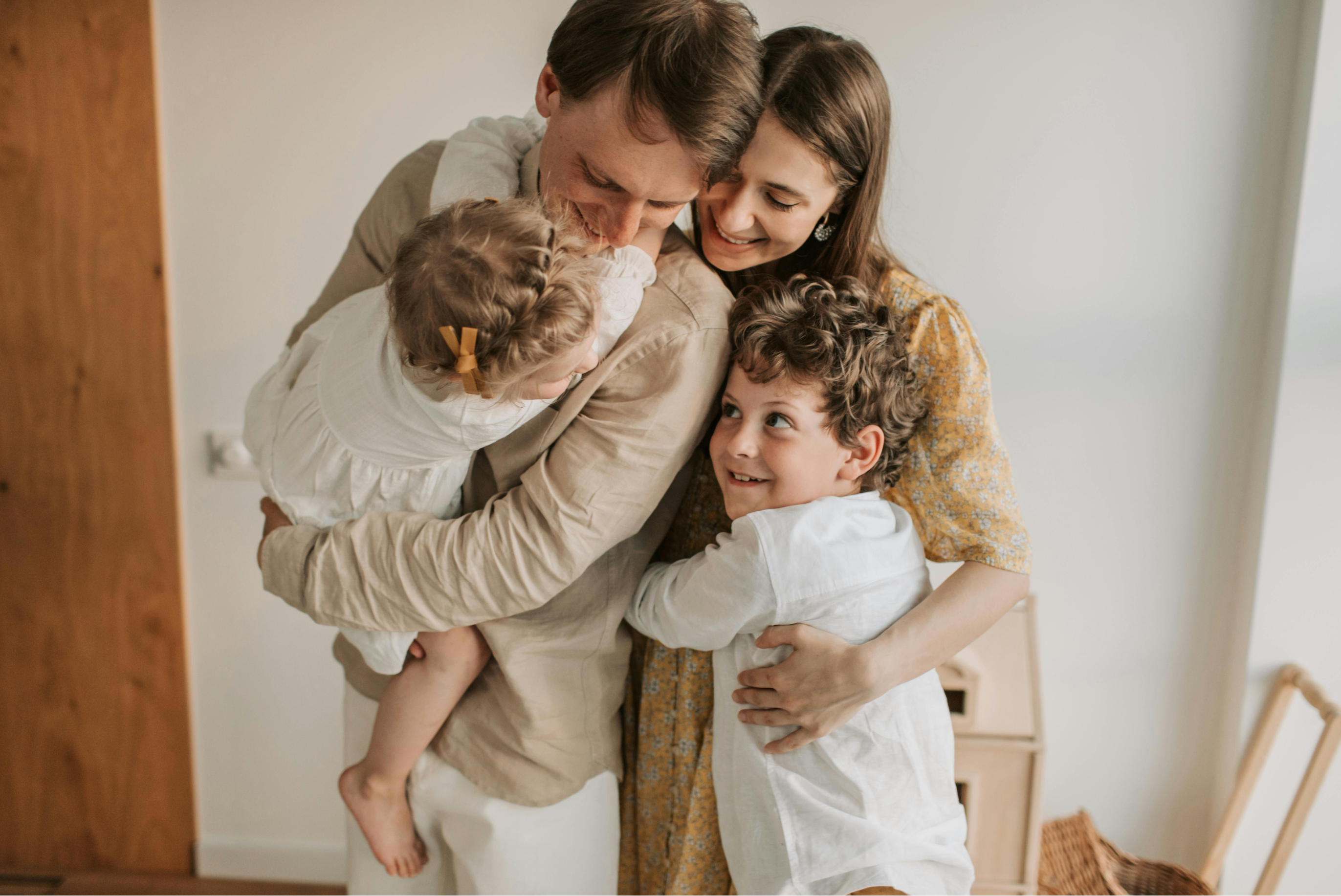 Family of four, two adults and two children, embracing each other indoors.