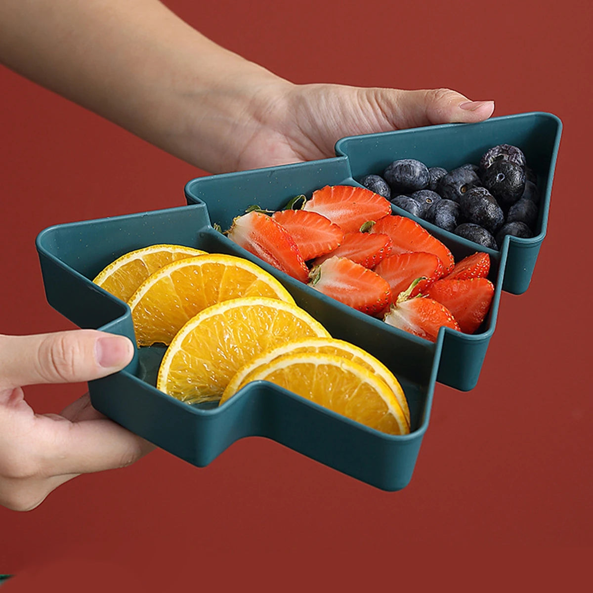Hand holding a blue compartmentalized container with sliced oranges and strawberries on a red background